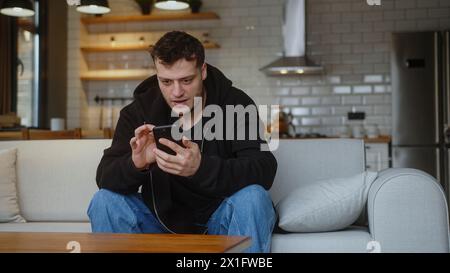 Jeune homme assis sur le canapé réagit à ce qui voit, surfer sur Internet sur le téléphone mobile, vérifier les e-mails, lire les nouvelles des médias, faire défiler les médias sociaux, en utilisant Mo Banque D'Images
