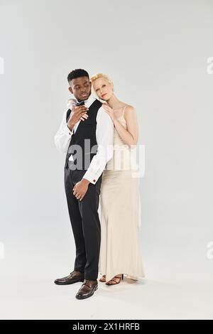 Une belle mariée blonde dans une robe de mariée et un marié afro-américain se tiennent côte à côte dans un studio sur un fond gris. Banque D'Images