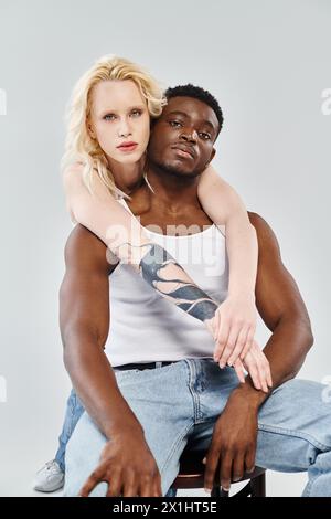 Un jeune couple interracial assis ensemble sur un tabouret dans un studio sur un fond gris. Banque D'Images