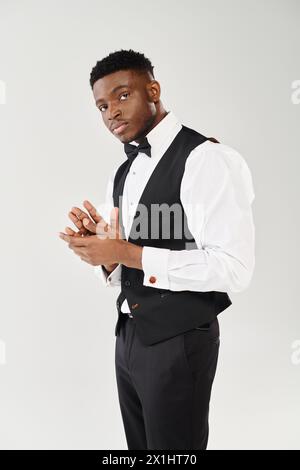 Un jeune homme afro-américain élégant se tient avec confiance dans un gilet noir et chemise blanche sur un fond gris studio. Banque D'Images