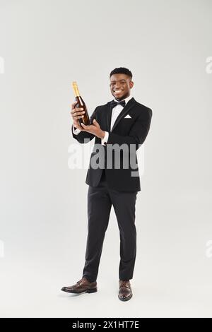 Beau marié afro-américain en smoking tient une bouteille de champagne, prêt à porter un toast de fête. Banque D'Images