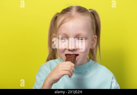 Une belle fille joyeuse aux cheveux blancs de sept ans mange une barre de chocolat sur fond jaune. Les enfants sont des amoureux de la malbouffe sucrée, horizonto Banque D'Images