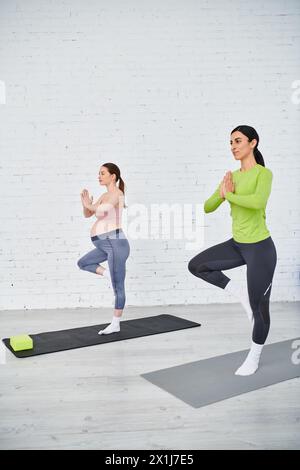 Deux femmes, dont une enceinte, posent gracieusement dans des positions de yoga, trouvant paix et force devant un mur de briques blanches. Banque D'Images