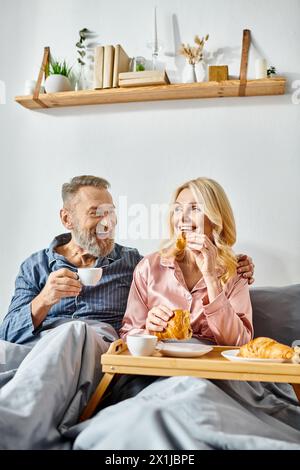 Un homme et une femme mûrs apprécient un repas assis sur un canapé confortable dans leur chambre confortable, vêtus de vêtements pour la maison. Banque D'Images