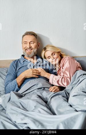 Un homme mature et une femme câlins dans le lit, portant des vêtements de maison confortables, appréciant les uns les autres compagnie dans leur chambre à coucher. Banque D'Images