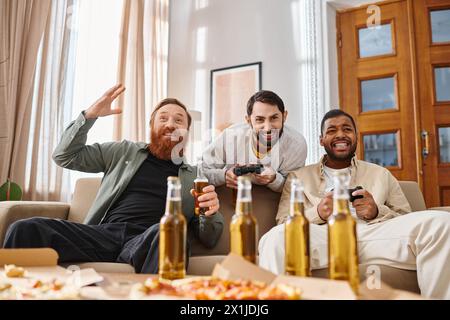 Trois hommes beaux et joyeux de différentes races se détendent sur un canapé avec de la bière et de la pizza, appréciant les uns les autres compagnie. Banque D'Images