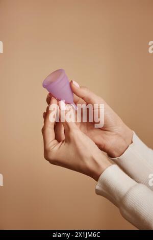 Gros plan minimal de mains féminines tenant la coupe menstruelle rose sur fond doux de couleur pêche Banque D'Images