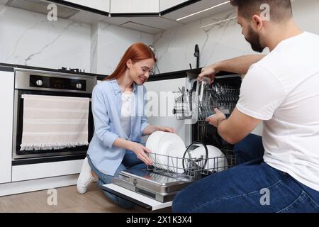Beau couple chargeant lave-vaisselle avec des assiettes dans la cuisine Banque D'Images