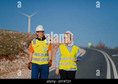 Une ingénieure avec une tablette et son collègue inspectent les éoliennes dans un champ, démontrant le travail d’équipe et le dévouement aux projets d’énergie renouvelable Banque D'Images