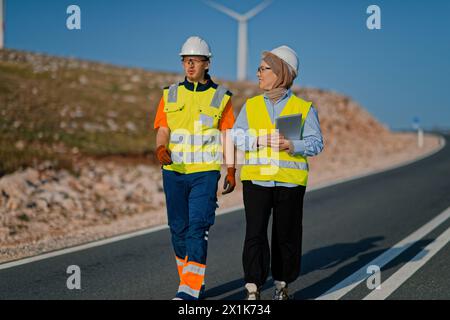 Une ingénieure avec une tablette et son collègue inspectent les éoliennes dans un champ, démontrant le travail d’équipe et le dévouement aux projets d’énergie renouvelable Banque D'Images