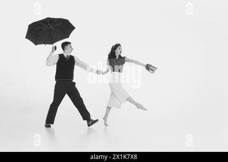 Charmant jeune couple, habillé en tenue vintage, se tenant la main et marchant joyeusement sous parapluie isolé sur fond blanc Banque D'Images