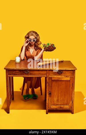 Femme élégante avec des lunettes de soleil tient hotdog dans le gant de baseball, avec chauve-souris au bureau en bois, sur fond de studio jaune. Banque D'Images