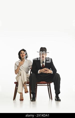 Élégant couple élégant, homme et femme en costumes classiques assis sur une chaise avec visage émotionnel et regarder l'émission, la télévision isolée sur fond blanc. Banque D'Images