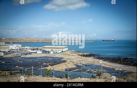 Panneaux solaires disposés sur les rochers dans le port de Luderitz en Namibie ; avec le vieux phare et Shark Island en arrière-plan. Banque D'Images