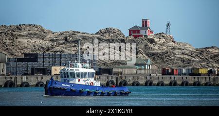 Le Pelican Tug passant devant le vieux bâtiment du phare sur l'île stérile et rocheuse des requins au port de Luderitz en Namibie. Banque D'Images