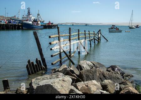 Vieille clôture brisée qui avance dans le port avec des navires et des bateaux amarrés et à l'ancre dans le port de Luderitz en Namibie. Banque D'Images