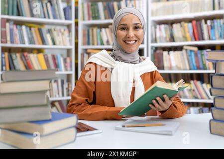 Une femme musulmane joyeuse en hijab est assise à un bureau entourée de piles de livres dans une bibliothèque, souriant tout en tenant un livre vert. Banque D'Images