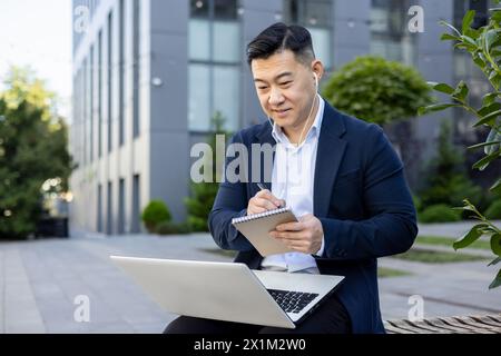 Jeune homme d'affaires asiatique travaillant à l'extérieur du centre de bureau, assis sur le banc portant des écouteurs, tenant un ordinateur portable et prenant des notes dans un ordinateur portable. Banque D'Images