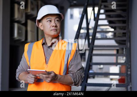 Photo d'un jeune homme asiatique ingénieur, architecte, contremaître debout dans un casque de sécurité et gilet à l'extérieur du bâtiment, tenant une tablette dans ses mains et regardant avec confiance sur le côté. Banque D'Images