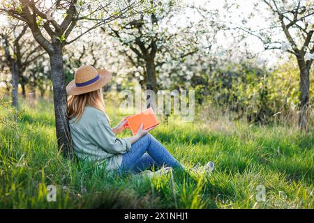 La femme se détendait dans le verger fleuri et lisait un livre pour améliorer sa pleine conscience et sa santé mentale. Détox numérique en extérieur au printemps Banque D'Images