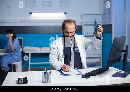 Un médecin spécialisé examine une image tomodensitométrique d'un patient tout en prenant des notes sur son presse-papiers. L'image montre un travailleur de la santé masculin caucasien examinant une radiographie thoracique d'une personne. Banque D'Images