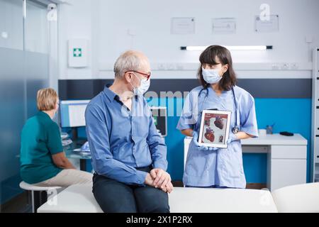 Homme senior regardant l'illustration de coeur sur tablette tenue par l'assistant médical. Praticienne décrivant le système cardiovasculaire et les traitements à un patient âgé de sexe masculin portant des masques. Banque D'Images