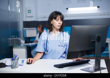 Image détaillée d'une infirmière caucasienne utilisant un ordinateur de bureau dans un cabinet hospitalier. Une praticienne portant des gommages bleus vérifie les dossiers médicaux sur un ordinateur dans une salle de clinique. Banque D'Images