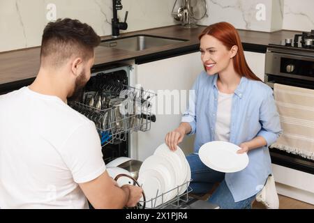 Beau couple chargeant lave-vaisselle avec des assiettes dans la cuisine Banque D'Images