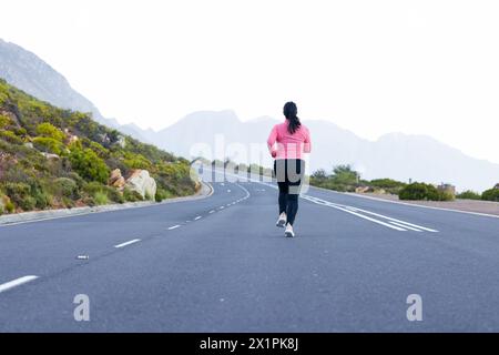 Randonneuse biraciale portant un haut rose et des leggings noirs, jogging sur une route avec espace copie Banque D'Images