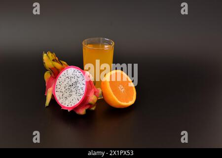 Un grand verre d'un mélange de jus de fruits tropicaux sur un fond noir, à côté des moitiés d'une orange mûre et pitaya. Gros plan. Banque D'Images