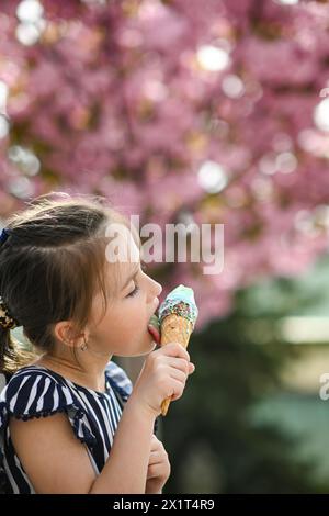 La petite fille lèche la crème glacée en été. Bel arbre rose fleuri dans le fond-bokeh. Banque D'Images