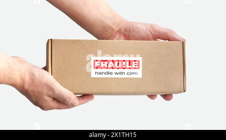 Une boîte en carton rectangulaire dans les mains pour l'emballage d'articles fragiles sur un fond clair Uni. Étiquette d'avertissement rouge, autocollant pour emballage « fragile » Banque D'Images