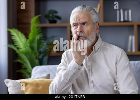 Un homme âgé inquiet dans une chemise décontractée est assis sur un canapé, touchant sa joue en raison d'un mal de dents, avec une expression douloureuse dans un cadre familial. Banque D'Images
