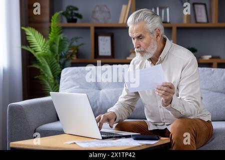 Homme âgé concentré avec les cheveux gris, travaillant sur un ordinateur portable tout en étant assis sur un canapé, examinant attentivement les documents dans un environnement confortable à la maison. Banque D'Images