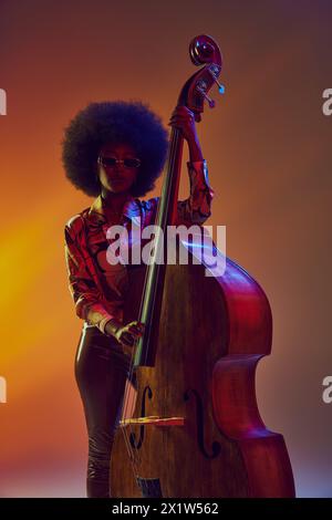 Musicienne afro-américaine confiante jouant la contrebasse dans la lumière néon sur fond dégradé. Banque D'Images