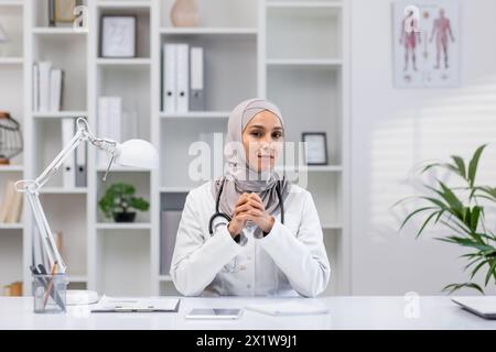 Portrait professionnel d’une médecin confiante portant un hijab, assise dans son bureau de clinique bien organisé, illustrant l’inclusivité et la diversité dans les soins de santé. Banque D'Images