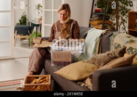 Jeune femme assise sur le canapé tout en choisissant et en pliant les vieux vêtements avant de les mettre dans la boîte et de les envoyer au centre de dons Banque D'Images