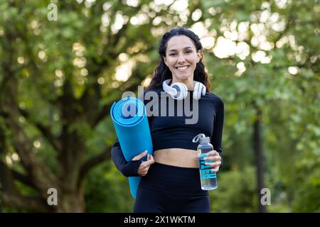 Une femme hispanique joyeuse tenant un tapis de yoga et une bouteille d'eau, prête pour une séance de fitness en plein air, avec des écouteurs autour du cou. Banque D'Images