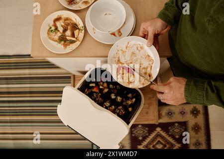 Vue de dessus des mains d'un jeune homme méconnaissable avec assiette jetant des restes de repas dans un seau en plastique ou une poubelle contenant de la terre Banque D'Images