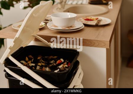 Seau en plastique ou poubelle avec couvercle ouvert contenant des restes d'aliments biologiques debout près du comptoir de cuisine avec de la vaisselle sale Banque D'Images