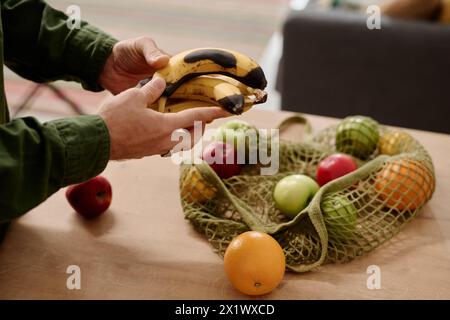 Mains d'un homme méconnaissable sélectionnant des fruits dans un sac à provisions tout en se tenant près du comptoir de la cuisine et tenant des bananes pourries Banque D'Images