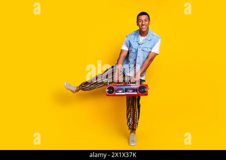 Photo pleine longueur de gars excentrique habillé jeans pantalon imprimé gilet dansant avec boombox dans les mains isolé sur fond de couleur jaune Banque D'Images