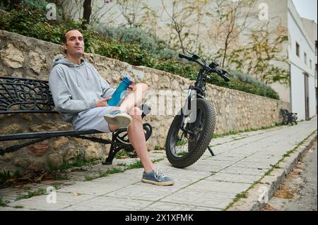 Jeune homme caucasien en sweat à capuche gris et short de sport, assis sur un banc près de son vélo électrique loué, se relaxant dans l'environnement urbain. Utilisation d'e-bik Banque D'Images