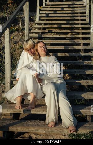 Deux sœurs partageant un moment tendre alors qu’elles s’embrassent sur les marches d’un escalier en bois altéré, baignées de la lueur chaude du soleil couchant. Banque D'Images