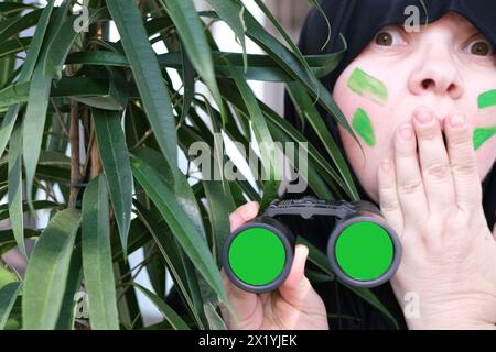gros plan curieuse femme islamique en peinture de guerre, tenant des jumelles de terrain avec zoom, des lentilles chromakey, regardant depuis les buissons, espionnant des husba infidèles Banque D'Images