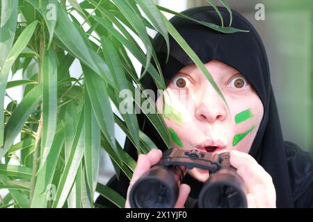 gros plan curieuse femme islamique en peinture de guerre, tenant des jumelles de champ avec zoom, regardant depuis les buissons, espionnant le mari infidèle, les voisins Banque D'Images