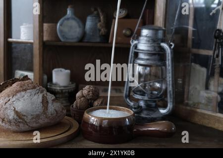 intérieur antique dans les tons bruns, bouteilles vintage, choses, lanterne antique, pain rond se trouve sur une planche à découper, le lait est versé dans un bol en argile dans un t Banque D'Images