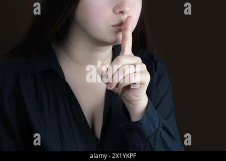 femme en gros plan, jeune fille dans une blouse noire aux cheveux longs pressée son doigt sur ses lèvres, encourage le silence, le geste sur un fond sombre, la dissimulation Banque D'Images