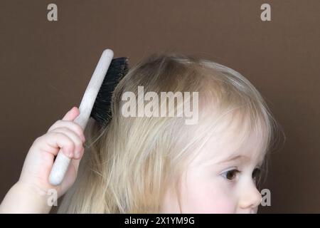 petite fille blonde se peignant indépendamment les cheveux avec une brosse en bois avec des poils naturels, enfant sur un fond brun foncé, concept d'hygiène, hai Banque D'Images