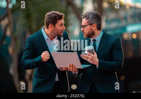 Deux hommes d'affaires surprisés utilisent un ordinateur portable à l'extérieur. Hommes d'affaires à la recherche ordinateur portable avec leur succès d'affaires dans le fond de la ville. Deux beaux jeunes Banque D'Images